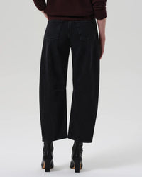 A person wearing Citizens of Humanity Miro Relaxed Jean in Raine, a high rise barrel leg washed black denim, paired with black heeled boots and a dark long-sleeve top. Shown from the back against a plain light gray background.