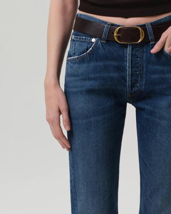 A person wearing Citizens of Humanity Winslow Low Rise Boyfriend jeans in Tinsley, styled with a black belt with a large oval gold buckle and a dark sleeveless top, shown from waist to mid-thigh with one hand by their side.