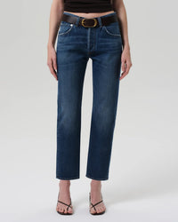 A person stands against a plain light gray background wearing Citizens of Humanity Winslow Low Rise Boyfriend in Tinsley jeans, paired with a black belt with a gold buckle, a dark top, and black heeled sandals.