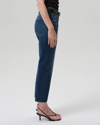 A person in profile wears blue Citizens of Humanity Winslow Low Rise Boyfriend jeans in Tinsley, made from regenerative cotton denim, paired with a black top and black strappy heeled sandals against a plain light gray background.