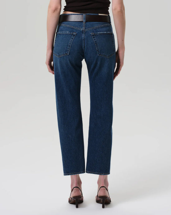 A person stands facing away from the camera against a plain light background, wearing Citizens of Humanity Winslow Low Rise Boyfriend jeans in Tinsley and brown heeled sandals.