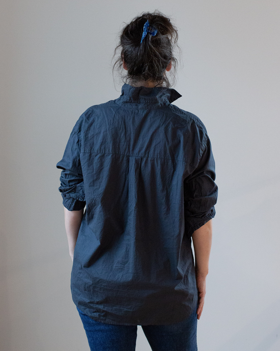 A person seen from behind wearing an oversized button-up shirt in CP Shades Ink Joss 1 Pocket Cotton Oxford and blue jeans with their hair tied up.