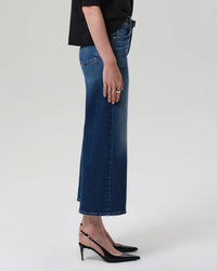 A person stands sideways against a plain light background wearing Citizens of Humanity Lyra Wide Leg Crop in Augusta jeans, a black top, and black slingback heels.