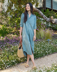 Woman in a Frank & Eileen Rory Maxi Shirtdress in Thyme Denim standing among flowering shrubs, holding a straw bag and smiling.