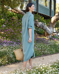 A woman in a long blue Frank & Eileen Rory Maxi Shirtdress in Thyme Denim carrying a straw basket, walking through a garden with blooming flowers.