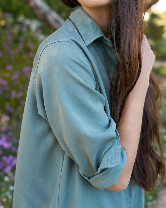 A woman in a Frank & Eileen Rory Maxi Shirtdress in Thyme Denim standing with her arm behind her head, partial view showing hair and sleeve.
