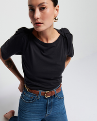 A young woman in a Nation LTD Deana Solid Envelope T Shirt in Jet Black and blue jeans, with a brown belt, looking intently at the camera. She has a tattoo on her left arm and is wearing gold.