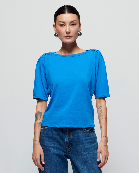 A woman with dark hair pulled back, wearing a Nation LTD Deana Solid Envelope T Shirt in Palace Blue and jeans, stands against a white background, showing visible tattoos on her arms.