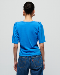 Woman seen from behind wearing a Nation LTD Deana Solid Envelope T Shirt in Palace Blue and denim jeans, with her hair styled in a bun and gold earrings visible.