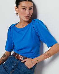 Woman in a Nation LTD Deana Solid Envelope T Shirt in Palace Blue with an envelope neckline and jeans with a brown belt, sporting tattoos on her arms, posing against a white background.