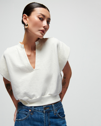 Woman with a sleek bun wearing a Nation LTD Lenon Crewneck Sweatshirt in Porcelain and denim jeans, showcasing gold earrings and tattooed arms, posed against a light background.