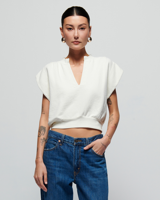 Woman in a white Nation LTD Lenon Crewneck Sweatshirt in Porcelain with a notch neckline and blue jeans, with tattoos on her arms, standing against a gray background.