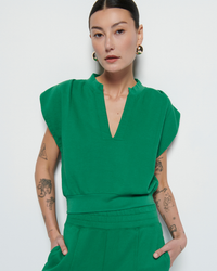 A woman in a Lenon Crewneck Sweatshirt in Verdant Green by Nation LTD, with short sleeves and a notch neckline, sporting various tattoos on her arms, poses against a white background.