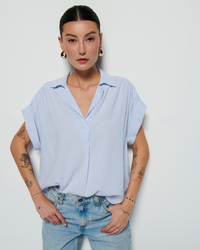 A woman with short hair wearing the Nation LTD Elliott Stripe Shirt in Mini Stripe and jeans, stands against a white background with visible tattoos on her arms.