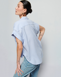 A woman with dark hair in a bun, wearing a Nation LTD Elliott Stripe Shirt in Mini Stripe and jeans, stands with her back to the camera, showcasing tattooed arms.