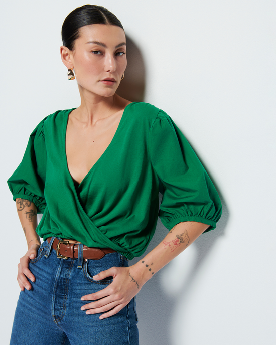 A woman in a Nation LTD Charlene Bubble Hem Top in Verdant Green and blue jeans posing with one hand on her hip against a white background.