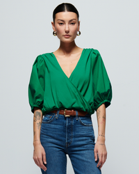 A woman with slicked-back hair wearing a green Nation LTD Charlene Bubble Hem Top in Verdant Green and jeans, standing confidently against a grey background.