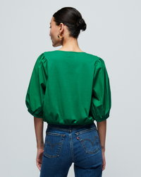 Woman standing with her back to the camera, wearing a green Nation LTD Charlene Bubble Hem Top in Verdant Green and blue jeans, against a gray background.