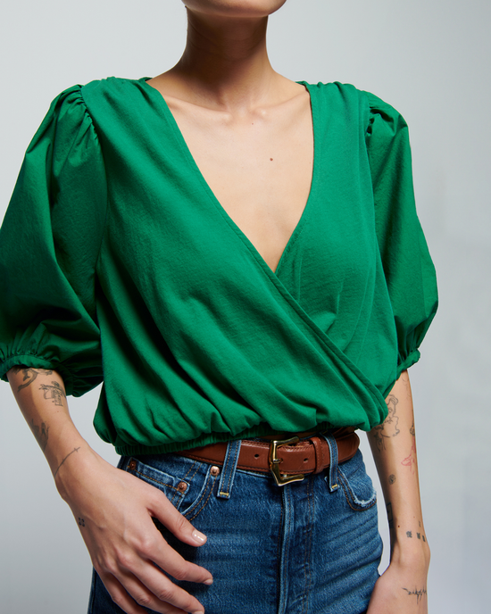 Woman wearing a green Nation LTD Charlene Bubble Hem Top in Verdant Green and blue jeans with a brown belt, focus on her outfit from the neck down.