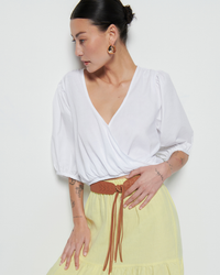 A woman in a white Nation LTD Charlene Bubble Hem Top and yellow skirt with a brown belt, looking away from the camera, showing her tattoos on one arm.
