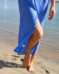 A person in a blue Frank & Eileen Rory Maxi Shirtdress walking barefoot on a sandy beach, with the focus on their lower body and the ocean in the background.