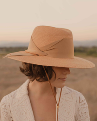 A woman in profile wearing a Field Gardenia in Tan fedora from Freya and a textured white jacket, standing outdoors with a soft-focus background of a dusky sky.
