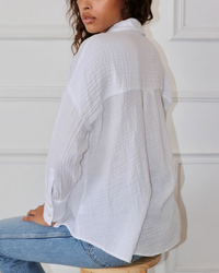 A woman wearing a Felicite Apparel Drop Shoulder Shirt in White and denim jeans, sitting sideways on a stool against a white door background.