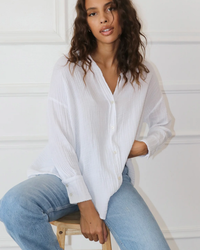 A woman sits on a stool, wearing a Felicite Apparel Drop Shoulder Shirt in White and blue jeans, with one hand resting on her knee, in a light-filled room.