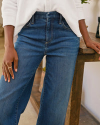Wearing Frank & Eileen's East Galway Long Wide Leg Pant in 1999 Blue Wash, a person stands indoors with one hand on a wooden table. Only their lower torso and upper legs are shown.
