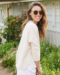 A smiling woman wearing sunglasses and a Frank & Eileen Anna Long Sleeve Capelet in Vintage White with a relaxed fit, standing in a garden.