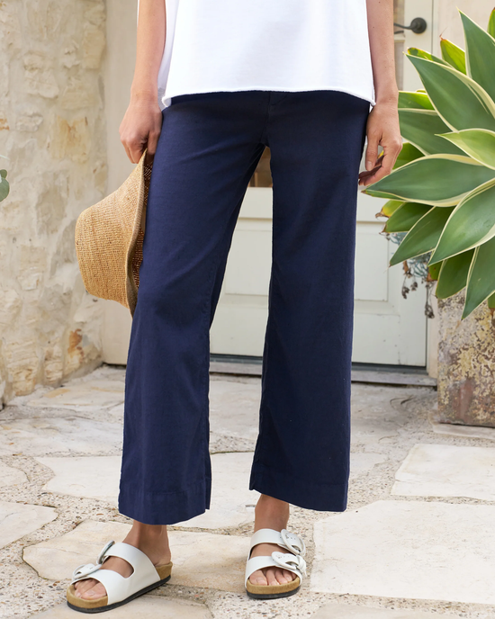 A person standing in a courtyard wearing Frank & Eileen's Wexford Trouser in Navy, a white t-shirt, white sandals, and holding a straw hat.