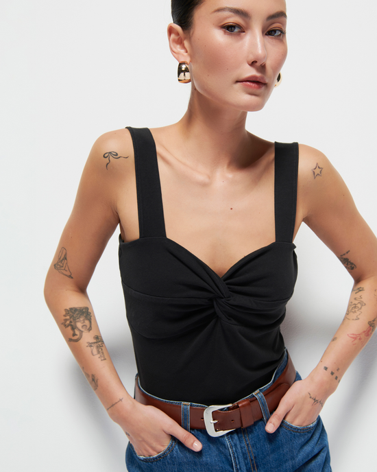 A woman wearing a black Nation LTD Mercy Twist Front Tank with a sweetheart neckline and jeans with a brown belt, showcasing multiple tattoos on her arms, poses against a white background.