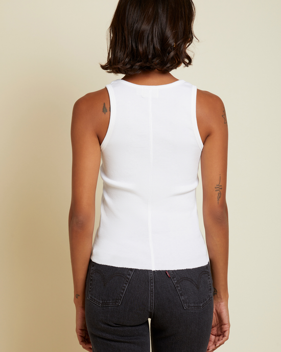 A woman stands facing away from the camera, wearing a Nation LTD Stella Tank in White with a scoop neckline and dark jeans, against a neutral background.
