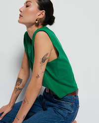A woman in a Nation LTD Collins Crewneck Solid Tank in Verdant Green and high rise denim jeans sits side-on, looking away from the camera, displaying multiple tattoos on her arms.