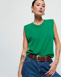 A woman in a Nation LTD Collins Crewneck Solid Tank in Verdant Green with high rise denim jeans, accessorized with a brown belt and hoop earrings, posing against a white background.