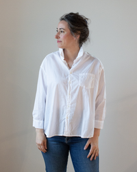 A woman wearing a CP Shades Joey Short Shirt in White Cotton Oxford with button-front closure and blue jeans, standing and looking to the side.