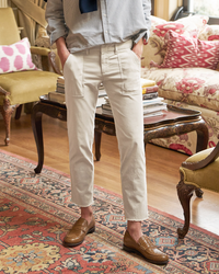 A person standing in a well-furnished room wearing Frank & Eileen Blackrock Utility Pants in Chalk, a tucked-in striped shirt with a dark tie, and brown woven loafers.