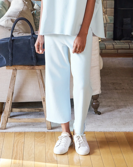 A person standing in a room wearing white sneakers, light blue trousers, and a matching top with a dark blue handbag on a wooden bench beside them. They are showcasing the Frank & Eileen Triple Fleece Catherine Cropped Wide Leg Sweatpant in Ice.