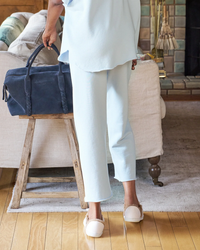 A person standing by a wooden stool holding a navy blue bag, wearing Frank & Eileen Catherine Cropped Wide Leg Sweatpants in Ice and white slip-on shoes.