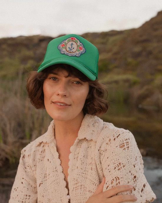 Woman with bob haircut wearing a lace cardigan and Freya's Sally Sells Trucker in Green, standing outdoors with a rocky landscape in the background.