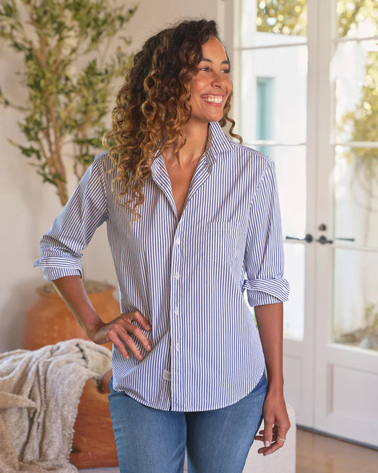 A smiling woman wearing the Frank & Eileen Eileen Relaxed Button Up Shirt in White & Navy Stripe stands indoors in blue jeans, one hand on her hip, with a plant and cozy decor in the background.