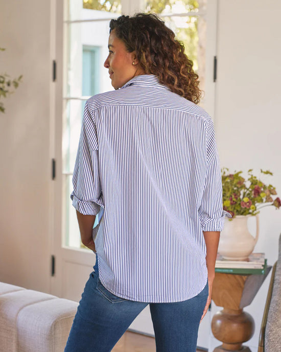 A woman with curly hair stands indoors, facing away, wearing the Frank & Eileen Eileen Relaxed Button Up Shirt in White & Navy Stripe with rolled-up sleeves and blue jeans. A table with a vase of flowers sits by a window in the background.