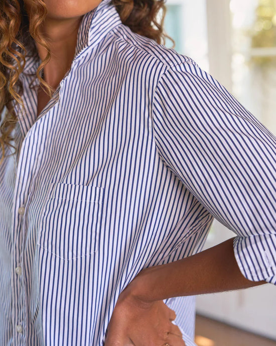 A person with curly hair wears the Frank & Eileen Eileen Relaxed Button Up Shirt in White & Navy Stripe, sleeves rolled up, standing indoors with their hand on their hip.