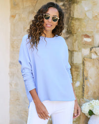 A woman in a Frank & Eileen Anna Long Sleeve Capelet in Saltwater and white pants, wearing sunglasses, stands confidently in front of a stone wall.