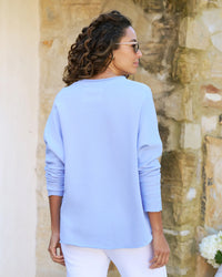 A woman wearing a Frank & Eileen Anna Long Sleeve Capelet in Saltwater and white pants, standing in front of a stone wall, seen from the back with her head turned to the side.
