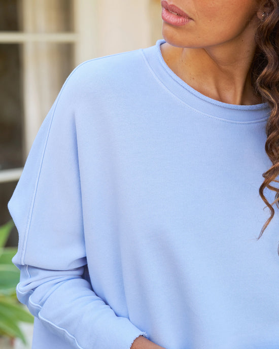 Close-up of a woman in a Frank & Eileen Anna Long Sleeve Capelet in Saltwater, focusing on the garment and part of her face, with a blurred background of greenery.