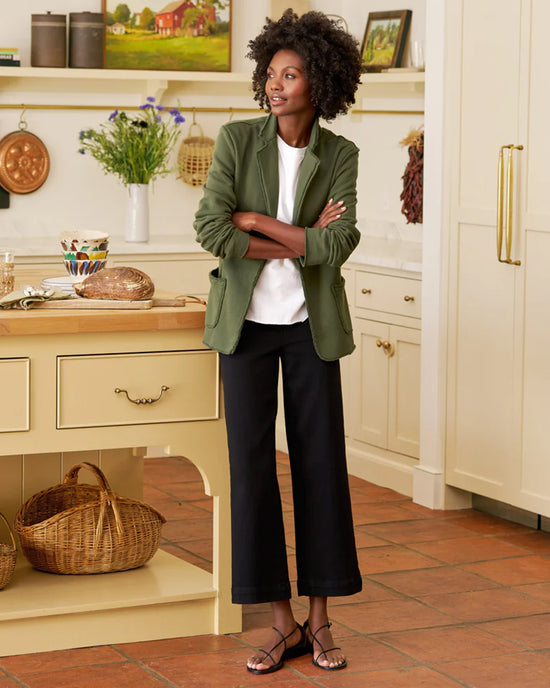 A woman in a green Frank & Eileen Dublin Knit Blazer in Caper, white top, and black wide-leg pants stands smiling with arms crossed in a cozy kitchen with cream cabinets, a central island, baskets, bread, and flowers.