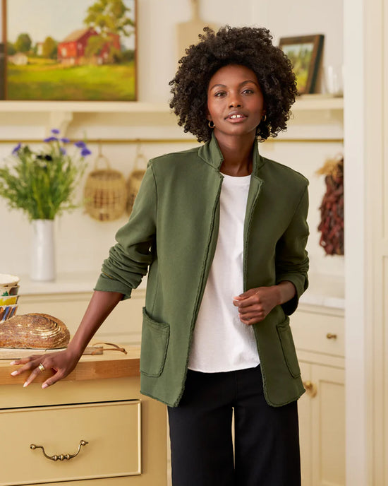 A woman with curly hair wears a Frank & Eileen Dublin Knit Blazer in Caper over a white shirt, standing in a cozy kitchen with cream cabinets. She rests a hand on a drawer beside bread, with flowers and baskets in the background.