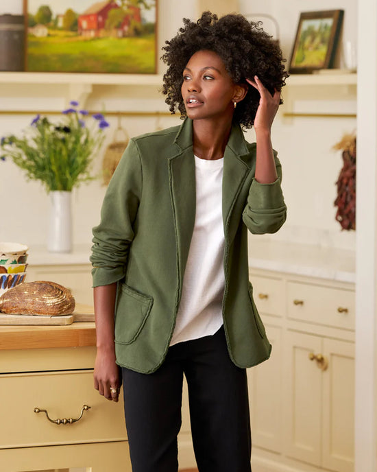 In a bright kitchen, a woman wearing a Frank & Eileen Dublin Knit Blazer in Caper and a white shirt stands touching her curly hair. Fresh bread, a mug, and flowers sit on the counter with light cabinets and shelves behind her.