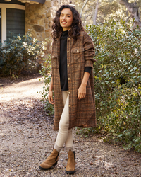 A woman stands on a garden path, smiling and modeling a Frank & Eileen Gavin Italian Wool Shirt Jacket in Orange & Brown Textured Plaid, light pants, and brown ankle boots.
